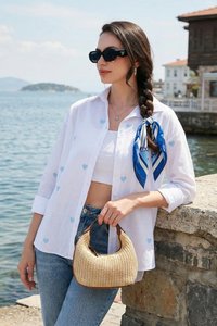 Femme portant des lunettes de soleil, chemise blanche avec des cœurs bleus, foulard bleu dans les cheveux tressés, tenant un sac à main tissé près d'une rambarde en pierre au bord de la mer.