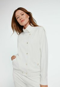 Biała, zapinana na guziki kurtka z wysokim kołnierzem, z złotymi guzikami i bocznymi kieszeniami. Gładka tekstura i luźny krój.