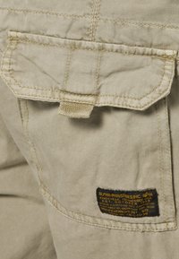 Beige cargo kalhoty se zadní kapsou, s klopou, šitými detaily a černou nálepkou značíci značku a specifikace.
