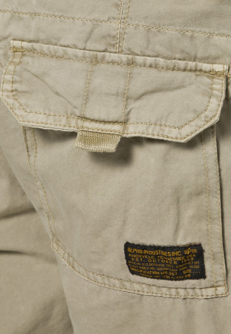 Beżowe spodnie cargo z tylną kieszenią, posiadające klapkę, wyszywane detale oraz czarną metkę z informacjami o marce i specyfikacjach.