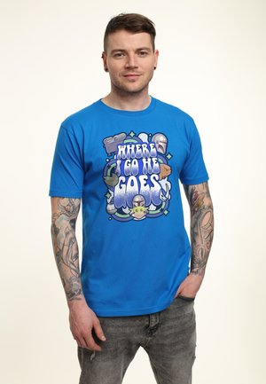 Young man with tattoos wearing blue T-shirt featuring Star Wars characters and text "Where I go he goes" in bold graphic design.