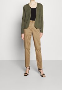 Person, der einen olivgrünen, strukturierten Cardigan, ein schwarzes Oberteil, beige, hochgeschnittene Hosen und schwarze, geschnürte High-Heels trägt.