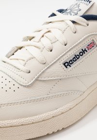 Zapatilla blanca de cuero con una suela intermedia texturizada, detalles perforados y acentos en azul. Incluye un logotipo de Reebok con la bandera de la Unión Jack.