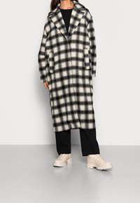 Dlouhý kostkovaný kabát v černobílé vlněné směsi, s oversize límcem, dvěma bočními kapsami a rovnými liniemi. Doplněno bežovými robustními botami.