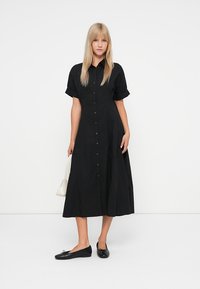 Abito nero con bottoni e maniche corte arrotolate, silhouette a trapezio e un tessuto leggero. Abbinato a ballerine nere e a una borsa bianca.