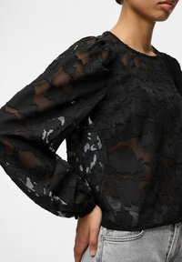 Melna caurspīdīga blūze ar ziedu izšuvumiem, garām uzpūstām piedurknēm un savāktiem aproču galiem. Kombinēta ar gaiši pelēnām džinsu biksēm, izceļot auduma faktūru un dizainu.