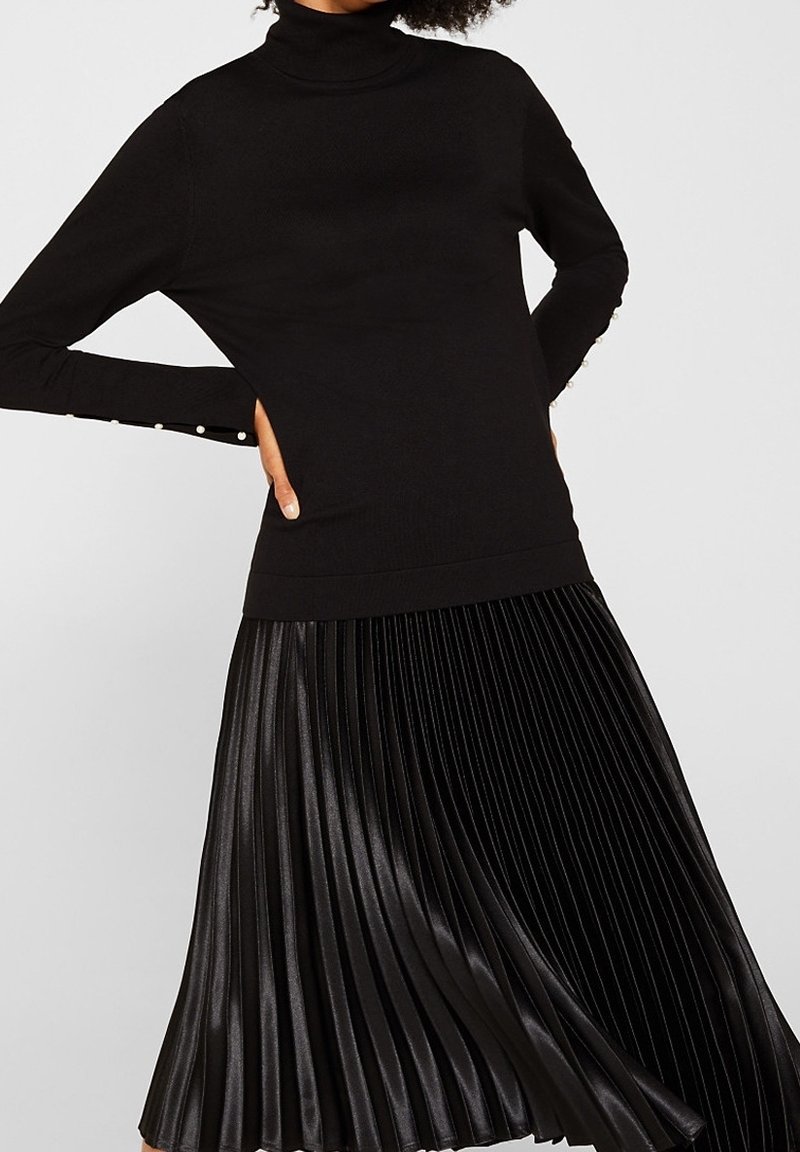 Černý rolákový svetr s dlouhými rukávy zdobený perlovými knoflíky. Kombinován s černou plisovanou sukní ke kolenům z lesklého materiálu.