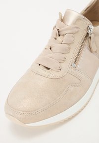 Baskets en suède beige avec une finition texturée, lacets plats, fermeture éclair sur le côté et semelle en caoutchouc avec des accents blancs.