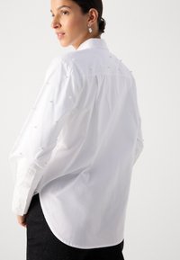 Camicia bianca con bottoni, caratterizzata da accenti in perle sulle spalle e sulle maniche. Vestibilità sartoriale con orlo curvo e design del colletto classico.