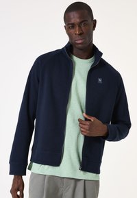 Marineblaue Zip-Jacke mit geripptem Material; verfügt über einen hohen Kragen und Logo-Detail auf der Brust. Getragen über einem hellgrünen T-Shirt.