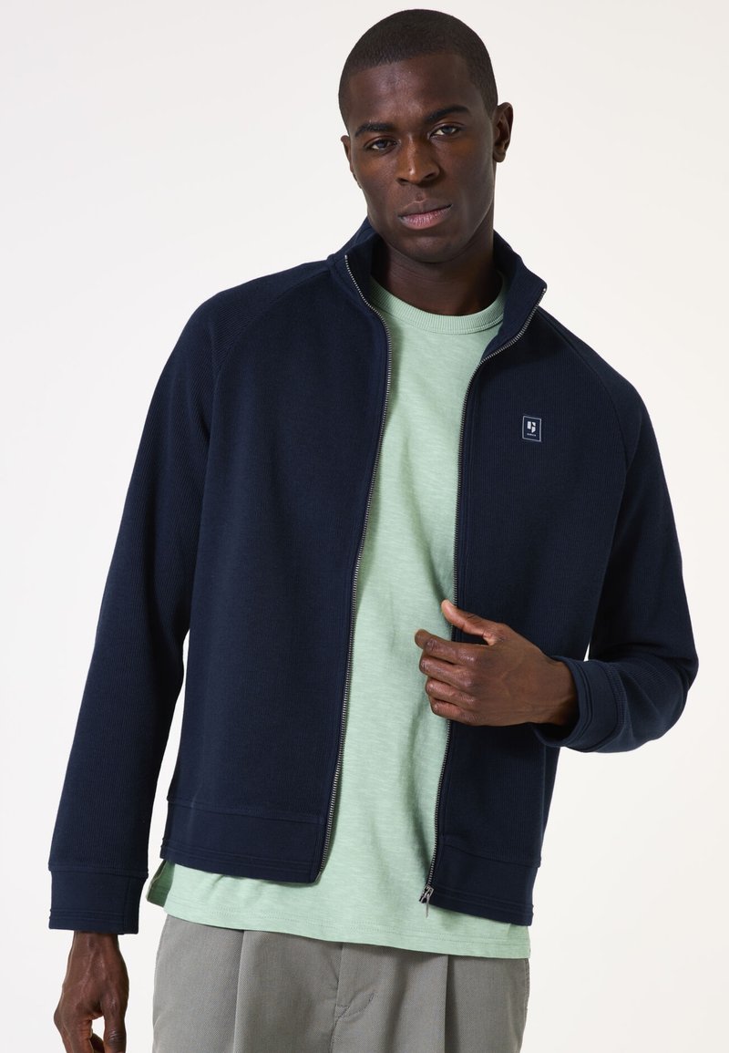 Marineblaue Zip-Jacke mit geripptem Material; verfügt über einen hohen Kragen und Logo-Detail auf der Brust. Getragen über einem hellgrünen T-Shirt.