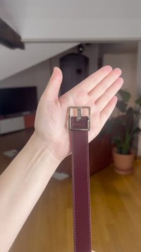 Ciemnoczerwony skórzany pasek z kwadratową, srebrną metalową klamrą; posiada widoczne przeszycia i gładką teksturę, trzymany na dłoni.