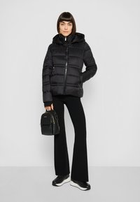 Zwarte pufferjas met capuchon, voorzien van een ritssluiting aan de voorkant, elastische manchetten en zijzakken. Gecombineerd met zwarte wijde broekspijpen en sneakers.