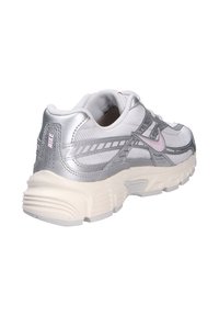 Zapatilla deportiva plateada y blanca con una parte superior de malla texturizada, detalles sintéticos lisos y una suela de goma gruesa. Presenta un detalle de logo en rosa.