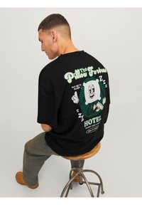 Černé tričko s oversized střihem, na kterém je vytištěna kreslená postavička polštáře a text na zádech, vytištěno v zelené a bílé barvě, zobrazující vtipné téma spánku.