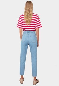 Žena s dlouhými vlnitými blond vlasy, oblečená v červeno-bíle pruhovaném tričku s krátkým rukávem, světlých modrých džínech s vysokým pasem a béžových botách, stojící zády.