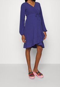 Fialové obepínací šaty s dlouhými rukávy a volánkovým lemem. V kombinaci s růžovými platformovými sandály s multibarevnými průhlednými pruhy.