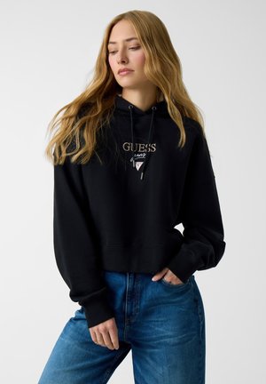 Felpa nera corta con coulisse e dettaglio logo dorato. Abbinata a jeans denim blu. Il tessuto appare morbido e strutturato.