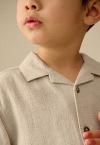 Licht beige overhemd met kraag, gemaakt van textuurrijk stof met een subtiel patroon. Het heeft een knoopsluiting en zichtbare stikdetails.