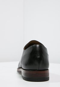 Chaussure habillée en cuir noir avec une finition lisse, un talon arrondi et une semelle en bois. Le design présente des lignes épurées et un detailing minimal.