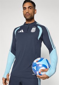 Άνδρας φορώντας γαλάζια και ανοιχτό γαλάζιο προπονητικό μπλουζάκι της ποδοσφαιρικής ομάδας Αργεντινής κρατώντας μια άσπρη και μπλε μπάλα ποδοσφαίρου Adidas στο ύψος της μέσης.