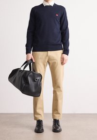 Marinblå tröja, khakifärgade byxor och svarta läderskor; håller en svart läderduffelväska med rundad form och korta handtag.