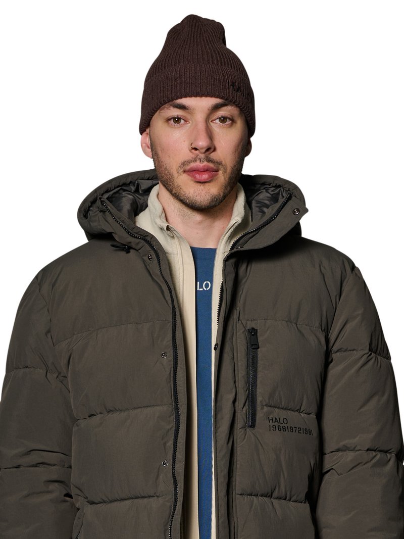 Mann trägt braune gestrickte Mütze, dunkel olivgrüne Steppjacke mit Reißverschluss und Druckknöpfen, beige Jacke und blaues Hemd darunter.