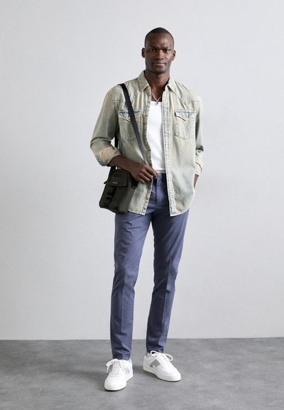 Camisa de mezclilla clara, camiseta blanca, pantalones azul marino, zapatillas blancas con detalles grises, sosteniendo un bolso cruzado verde oscuro. Diseño simple y funcional.