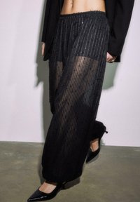 Crne prozirne hlače s teksturiranim dizajnom s okomitim ukrasima nalik na perlice. Elastični pojas i suženi rub, uparen s crnim štiklama.