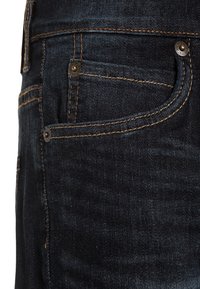 Tummansiniset farkut, joissa lähikuva etutaskusta. Ominaisuuksina kontrastisauma ja metallinappi, jotka korostavat sileää pintaa.