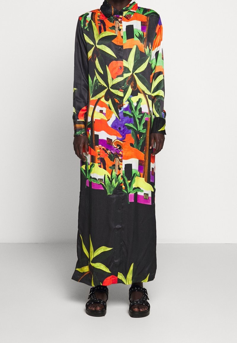 Robe longue boutonnée avec des motifs abstraits colorés de plantes tropicales et de maisons, portée avec des sandales noires cloutées sur un fond neutre.