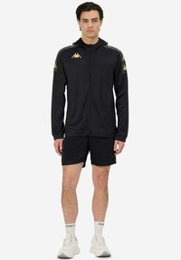 Giacca sportiva nera con zip frontale, caratterizzata da accenti in oro sulle spalle, abbinata a pantaloni corti neri e scarpe da ginnastica bianche.