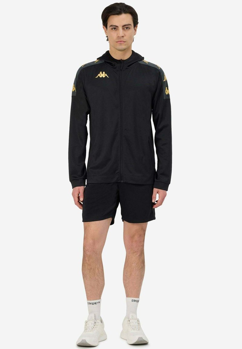 Giacca sportiva nera con zip frontale, caratterizzata da accenti in oro sulle spalle, abbinata a pantaloni corti neri e scarpe da ginnastica bianche.