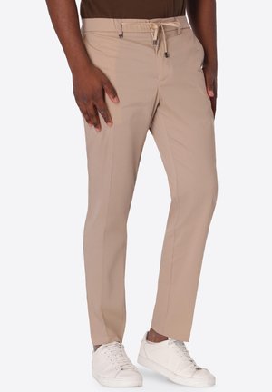 Uomo con pantaloni beige slim fit con coulisse in vita e sneaker bianche, in piedi con le mani rilassate lungo i fianchi.