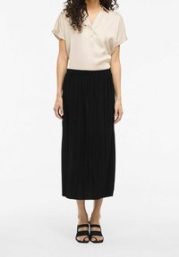 Blusa beige a maniche corte con bottoni, abbinata a una gonna midi plissettata nera e sandali neri con cinturini, dotata di una vita elastica arricciata.