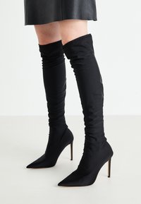 Botas negras por encima de la rodilla hechas de tela suave, con punta afilada y alto tacón de aguja; diseño fruncido en la parte superior.