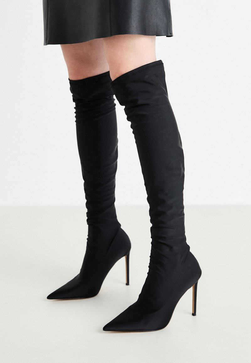 Botas negras por encima de la rodilla hechas de tela suave, con punta afilada y alto tacón de aguja; diseño fruncido en la parte superior.