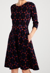 Kvinna i en knälång, långärmad klänning med ett röd- och marinblått geometriskt mönster, med höger hand i fickan.