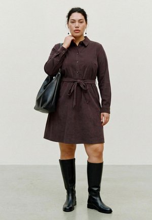 Une femme porte une robe-chemise marron foncé ceinturée, des bottes noires jusqu’aux genoux et un grand sac noir porté à l’épaule.