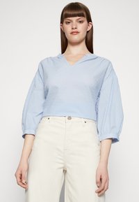 Blusa cropped azul-claro com decote em V, tecido com bolinhas texturizadas e mangas bufantes elásticas. Combinada com jeans de cintura alta off-white.