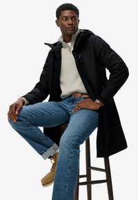 Zwarte waterdichte jas met een capuchon, over een grijze trui. Blauwe jeans met opgerolde broekspijpen en beige schoenen. Man zit op een houten kruk.