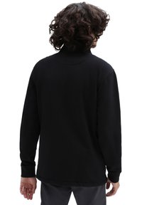 Persona con capelli ricci che indossa una maglietta nera a maniche lunghe e pantaloni scuri, vista da dietro su uno sfondo bianco.