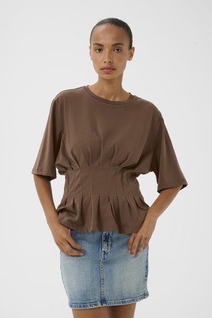 Kvinde står med fronten mod kameraet iført en brun top med samlet talje og korte ærmer samt en lyseblå denimnederdel mod en ensfarvet baggrund.