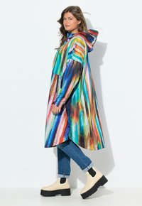 Femme portant un manteau à capuche rayé multicolore, un jean bleu retroussé et des bottes compensées beiges, se tenant devant un fond blanc.