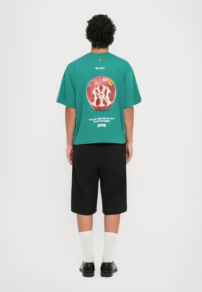 Green short-sleeve T-shirt with graphic of an apple and text on the back. Paired with black shorts, white socks, and black shoes.