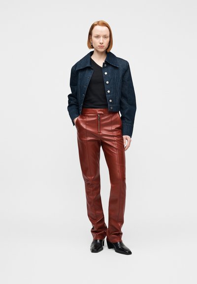 Νέα γυναίκα με κοντά κόκκινα μαλλιά που φοράει σκούρο τζιν μπουφάν, μαύρο τοπ, καφέ δερμάτινο παντελόνι ψηλής μέσης και μαύρες μπότες με τακούνι, στέκεται μπροστά σε γκρίζο φόντο.