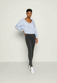 Sweater oversized azul claro com decote em V e mangas balão, combinado com jeans skinny cinza escuro e ténis brancos, de pé contra um fundo neutro.