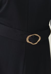 Ceinture en tissu noir avec une boucle ovale en or texturée et plusieurs trous. Design simple axé sur la fonctionnalité et le détail.