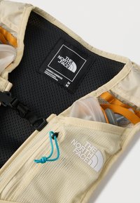 Κοντινή λήψη μπεζ γιλέκου The North Face με μαύρο εσωτερικό από δίχτυ, μπλε τράβηγμα φερμουάρ και διαφανές μπουκάλι νερού στην μπροστινή τσέπη.