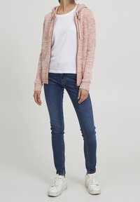 En vuxen som bär en rosa stickad zip-up hoodie, t-shirt i vitt, blå skinny jeans och vita sneakers, står mot en neutral bakgrund.